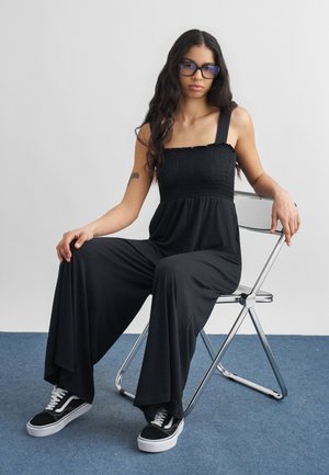 Fiatal nő hosszú sötét hajjal, fekete szemüveget visel, ujjatlan fekete overallt és fekete sportcipőt visel, fém összecsukható széken ül.