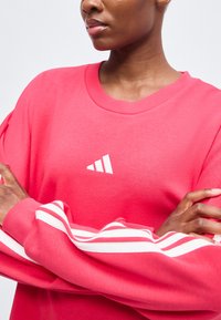 Άτομο που φοράει ένα φωτεινό ροζ φούτερ Adidas με λευκές ρίγες στα μανίκια, σταυρώνει τα χέρια μπροστά σε απλό φόντο.