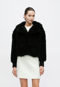 Μαύρο τζάκετ από ψεύτικη γούνα με φαρδύ γιακά, κομψή κοντή σχεδίαση και υφή. Φορεμένο πάνω από λευκό φόρεμα με υφή.