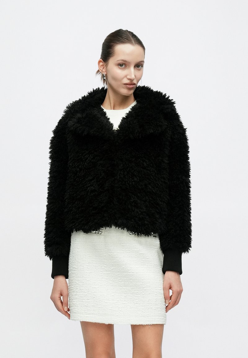 Μαύρο τζάκετ από ψεύτικη γούνα με φαρδύ γιακά, κομψή κοντή σχεδίαση και υφή. Φορεμένο πάνω από λευκό φόρεμα με υφή.