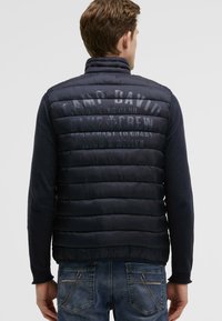 Gilet imbottito blu navy con colletto alto, design trapuntato e logo in rilievo sul retro. Indossato sopra una maglietta a maniche lunghe scura e jeans blu.
