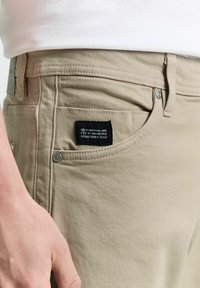 Beige színű, finom textúrájú pamut nadrág, fekete márkacímmel, fémből készült gombos záródással és oldalsó zsebbel, melyen látható varrás található.