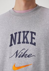 Gros plan d’un sweat-shirt gris avec le mot « NIKE » en gras, « Nike » en écriture manuscrite en dessous, et un logo Nike swoosh orange brodé en dessous.