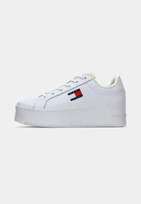 Baskets à plateforme blanches avec une tige synthétique lisse, un bout rond, une fermeture à lacets, et un patch logo avec des accents rouges et bleus sur le côté.