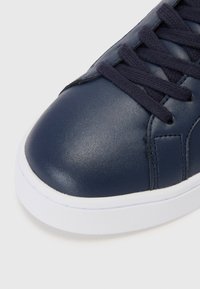 Tumedast sinine nahkse sneaker, millel on ümar nina, lame valge tald ja tumedad paelad, koos tekstuursete paneelide ja minimaalsete õmblusteenustega.