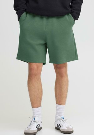 Person iført grønne knælange shorts, sort top, hvide sokker og hvide sneakers med sorte striber, stående med hænderne i lommerne.