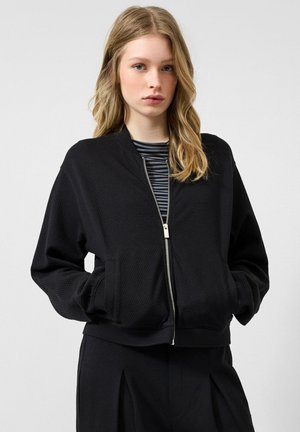 Junge Frau mit blondem Haar, die eine schwarze, strukturierte Reißverschlussjacke über einem gestreiften Hemd trägt, Hände in den Taschen, vor einem schlichten Hintergrund.