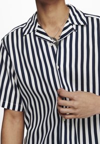 Persona che indossa una camicia a righe verticali navy e bianche a maniche corte, tenendo il tessuto vicino al centro del petto, rivelando una collana d'argento.