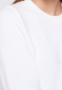 Weißer Pullover mit strukturiertem gestreiften Design, Rundhalsausschnitt und weichem Strickstoff. Fokus auf einer Nahaufnahme des Ärmelbereichs.