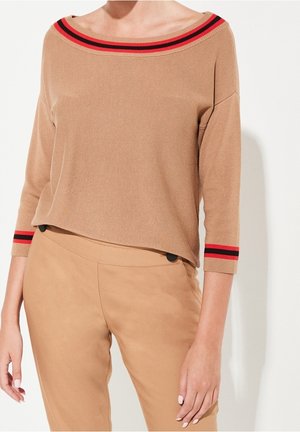 Kobieta ubrana w beżowy dzianinowy top z czerwonymi i czarnymi paskami na dekolcie i rękawach, w połączeniu z pasującymi beżowymi spodniami.