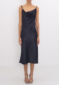Satin midi dress in navy blue with a cowl neckline, thin straps, and a fluid silhouette. Pair of gold-accented sandals visible.