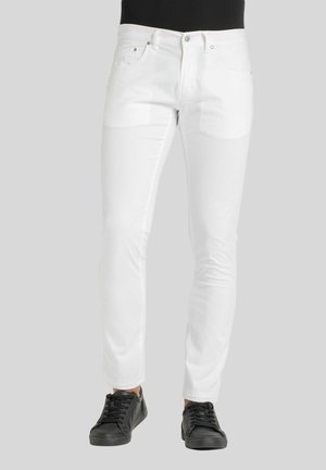 Jean en denim blanc avec une coupe ajustée, présentant des poches avant, une fermeture éclair et une texture lisse, légèrement brillante. Associés à des baskets noires.