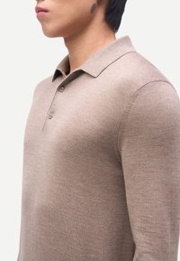 Junger Mann trägt ein beiges, langärmeliges Strick-Poloshirt mit zwei Knöpfen, dargestellt von der Brust aufwärts, blickt nach links vor weißem Hintergrund.