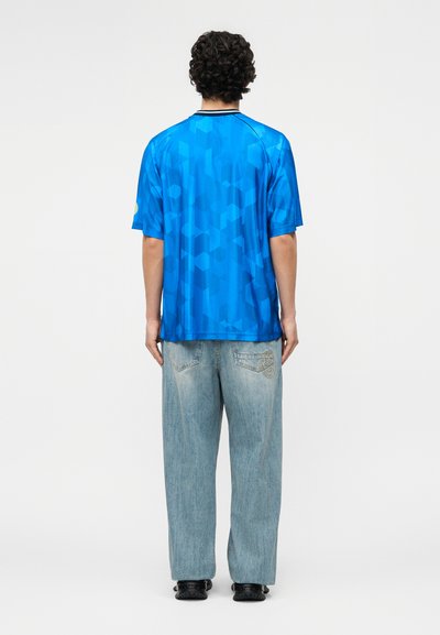 Άνδρας με σκούρα σγουρά μαλλιά στέκεται πλάτη, φορώντας μπλε πουκάμισο με γεωμετρικά σχέδια και κοντά μανίκια, καθώς και ανοιχτόχρωμο τζιν παντελόνι με μαύρα παπούτσια.