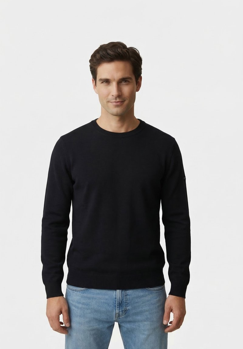 Uomo di fronte che indossa un maglione nero girocollo e jeans azzurri chiari su uno sfondo semplice e chiaro.