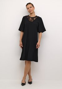 Schwarzes knielanges Kleid mit einer Spitzenakzentuierung am Ausschnitt. Der Stoff hat eine gerippte Textur und breite Ärmel. Getragen mit schwarzen High Heels.