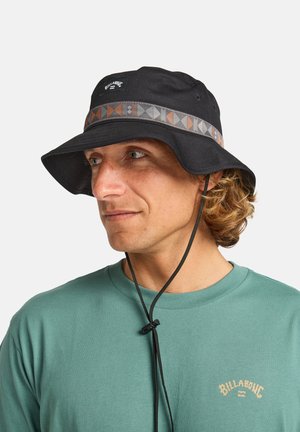 Man with curly hair wears a black bucket hat featuring a patterned band and a teal shirt with a logo, looking slightly to the side.