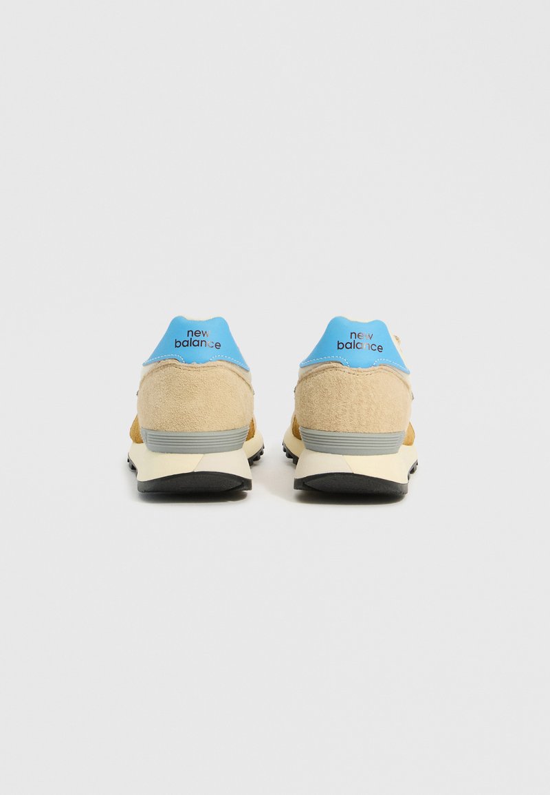 Sandfarbene und blaue Sneaker mit einem Obermaterial aus Wildleder, kontrastierenden hellbeigen Akzenten, einem gepolsterten Schaft und schwarzen Gummisohlen. "New Balance"-Logo an der Ferse.