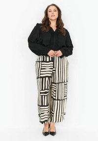 Donna che indossa una blusa nera a maniche lunghe con pieghe e pantaloni cremosi a gamba larga con audaci strisce geometriche nere, in piedi con le mani unite.