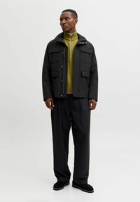 Čierna vodeodolná bunda s viacerými vreckami, skombinovaná so zeleným pleteným svetrom. Nohavice sú široké a topánky sú tmavé a štruktúrované.