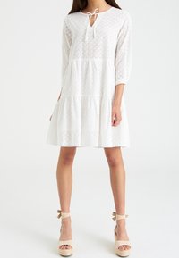 Vestido blanco de algodón con mangas de tres cuartos, patrón de ojal, diseño en capas y escote en forma de llave con detalle de lazo. Llevado con cuñas de esparto.