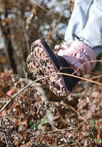 Lichtroze geïsoleerde laars met een gestructureerde zwarte zool, voorzien van een robuust profiel. De laars is gedeeltelijk ondergedompeld in gebladerte.