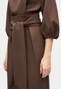 Κοντινό πλάνο προσώπου που φοράει καφέ φόρεμα με πλατιά ζώνη στη μέση και μαζεμένα μανίκια τριών τετάρτων, πάνω σε απλό ανοιχτό φόντο.