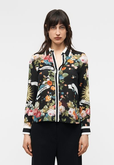 Γυναίκα που φοράει μαύρη μπλούζα με floral σχέδιο και λευκά τελειώματα, διακοσμημένη με ήλιο, λουλούδια, πουλιά και κείμενο, συνδυασμένη με μαύρο παντελόνι, στέκεται σε λευκό φόντο.