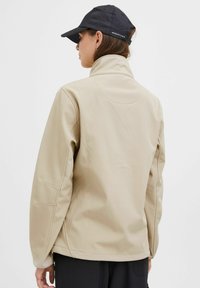 Beige softshell jacket with a high collar, designed with a smooth texture and understated seam details. Pairing with a black cap.