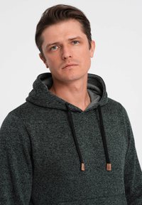 Felpa con cappuccio verde scuro, foderata in grigio, con accenti in cordino e tessuto testurizzato. Presenta una tasca a marsupio e un branding minimalista.