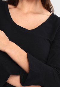 Schwarzes V-Ausschnitt-Oberteil mit langen Ärmeln und ausgestellten Manschetten, aus einem weichen, glatten Stoff gefertigt. Hat eine enganliegende Form und minimale Designdetails.