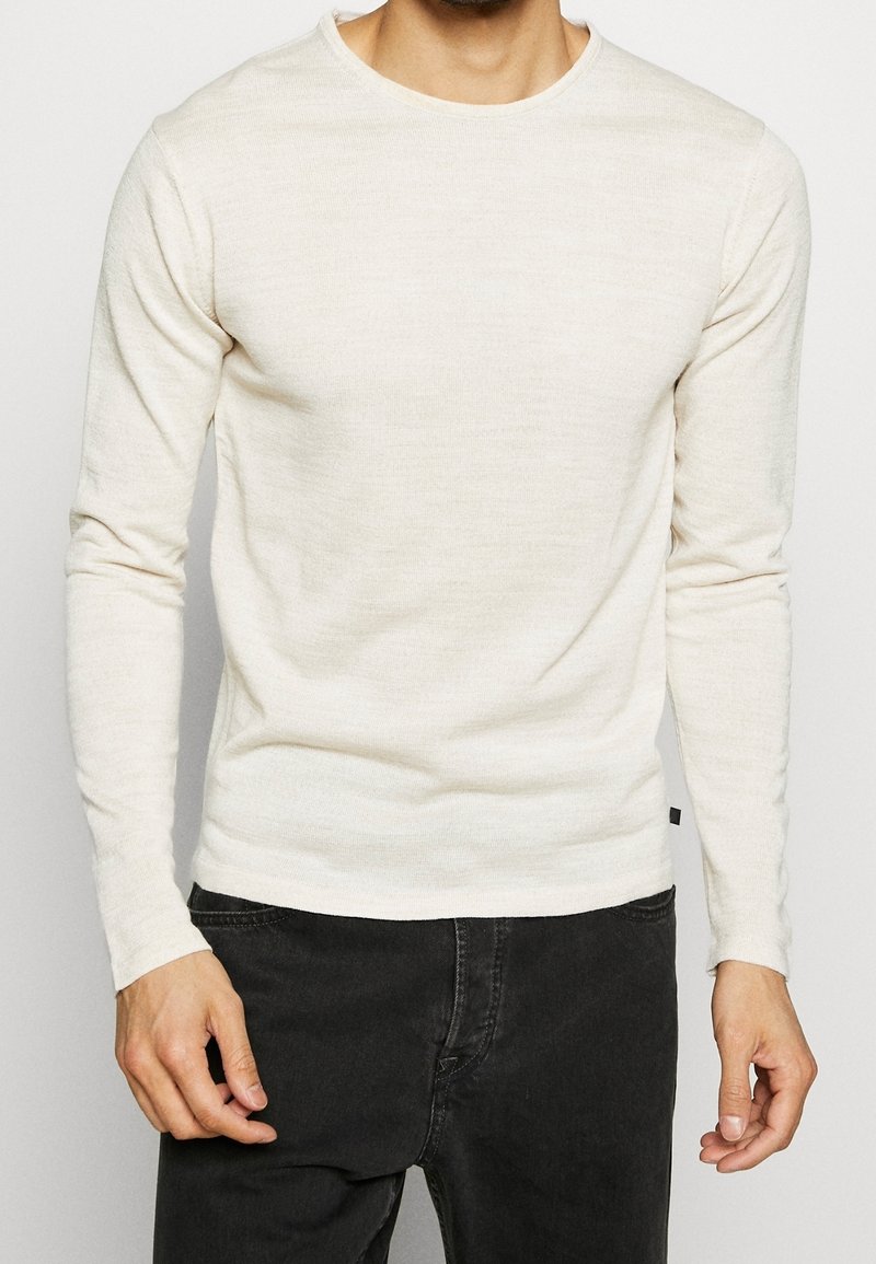 Homme portant un pull beige uni à manches longues associé à un jean noir, debout devant un fond neutre.
