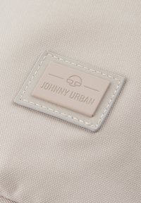 Tessuto beige chiaro con una superficie testurizzata, caratterizzato da un'etichetta rettangolare in rilievo beige che reca la scritta "JOHNNY URBAN," bordata da cuciture a contrasto.