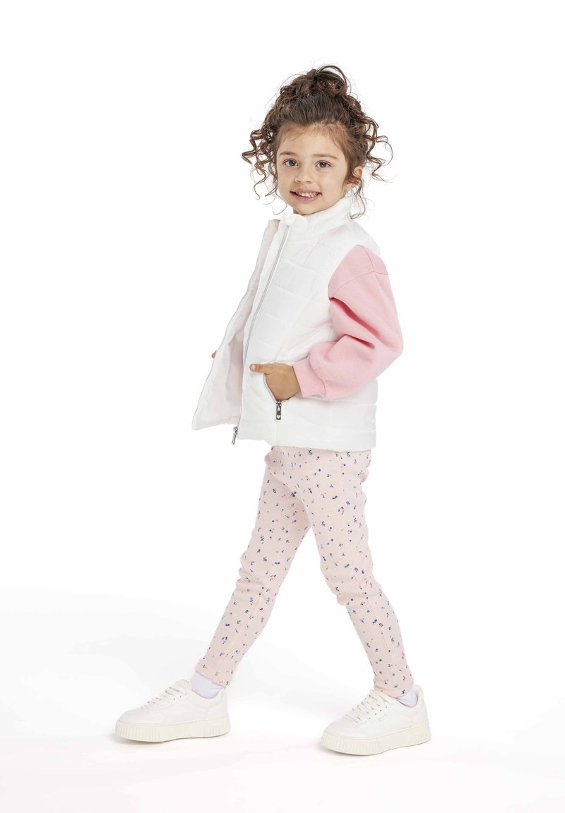 Vit vadderad väst över en rosa långärmad skjorta, matchad med rosa mönstrade leggings och vita sneakers. Har en dragkedja fram och fickor.