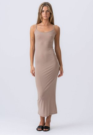 Nő, aki előre nézve áll, ujjatlan, testhezálló bézs maxi ruhát és fekete nyitott orrú papucsot visel, egyszerű világos háttér előtt.