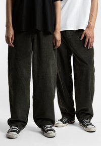 To personer står med hendene på sidene, iført løse, grønne kordfløyelsbukser og svarte kanvas sneakers. Stoffet har en vertikal ribbet tekstur.