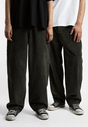 Twee personen staan met hun handen langs hun zij, gekleed in losse, groene corduroy broeken en zwarte canvas sneakers. De stof heeft een verticale ribbelstructuur.