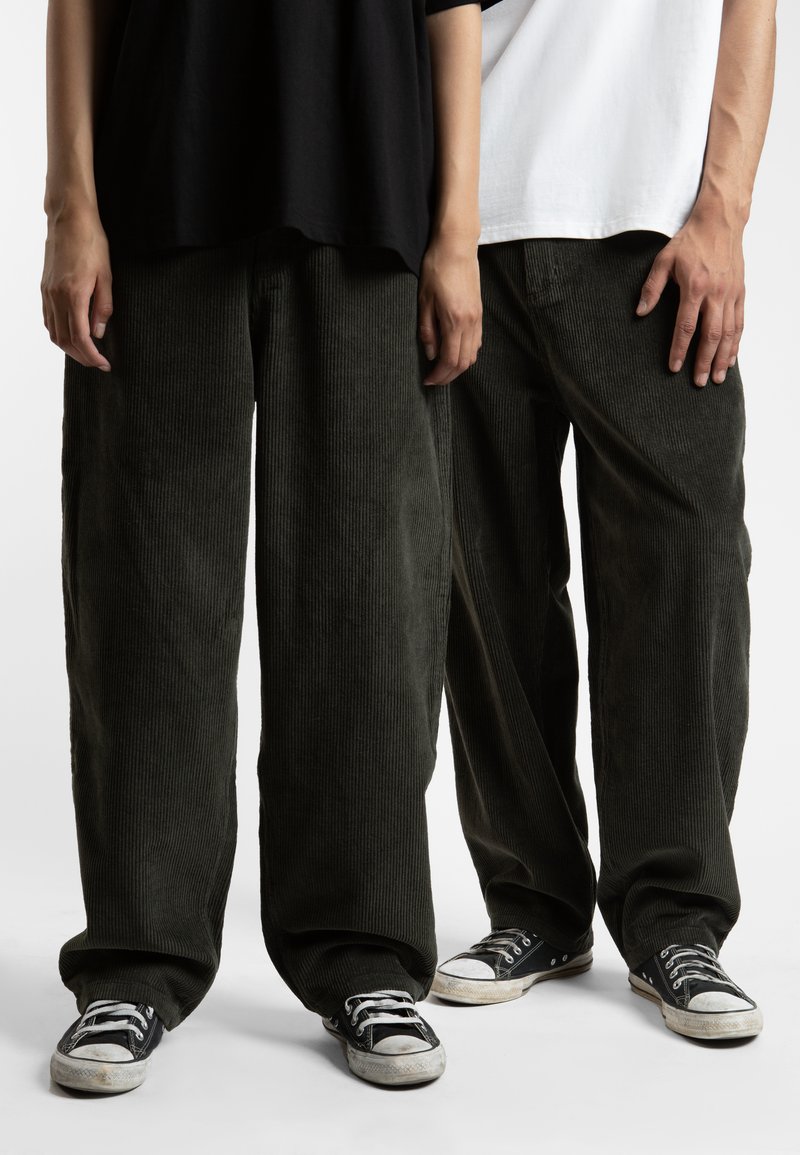 To personer står med hendene på sidene, iført løse, grønne kordfløyelsbukser og svarte kanvas sneakers. Stoffet har en vertikal ribbet tekstur.