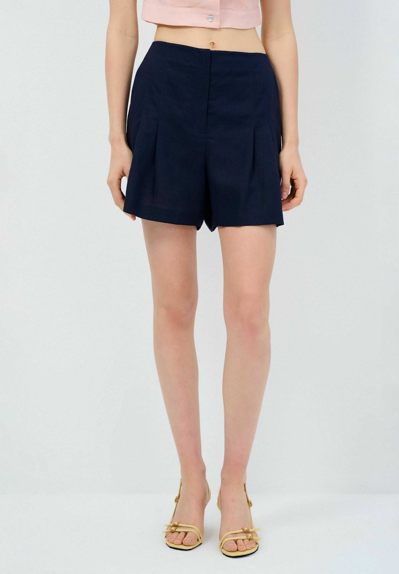 Pantalones cortos de marinero con pliegues, hechos de una tela ligera, que presentan una cintura limpia y un ajuste entallado, combinados con sandalias de color claro con tiras.