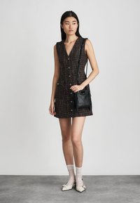 Ärmelloses Kleid aus dunkelbraunem Tweed mit bunten Fäden, mit einer Knopfleiste vorne. Kombiniert mit einer kleinen schwarzen Lederhandtasche.