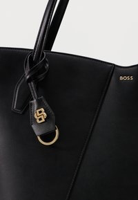 Sac fourre-tout en cuir noir avec poignées nouées, logo "BOSS" doré, et emblème doré avec initiales suspendu à une poignée.