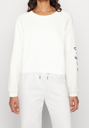 Kvinde iført en enkel hvid langærmet sweatshirt og hvide bukser, sweatshirt har delvist synlig sort grafisk tekst på højre ærme.