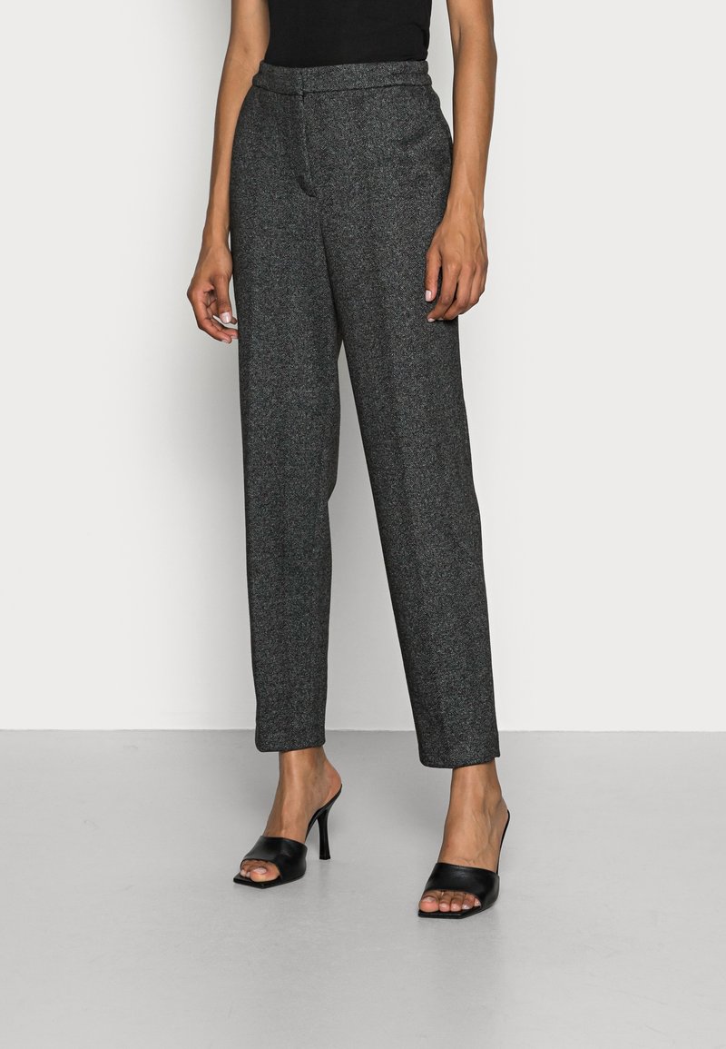 Personne portant un pantalon gris foncé ajusté et des sandales noires à talons hauts et à bouts ouverts sur un fond clair uni.