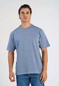 Jasnoniebieska koszulka z krótkim rękawem, uszyta z gładkiego materiału, z okrągłym dekoltem, luźnym krojem i prostym dołem; zestawiona z niebieskimi dżinsami.