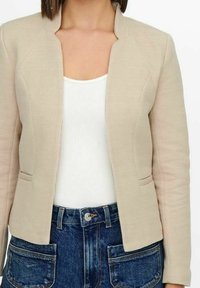 Blazer beige à texture chevron, design ouvert à l'avant et manches longues. Associé à un haut blanc côtelé et un jean en denim foncé.
