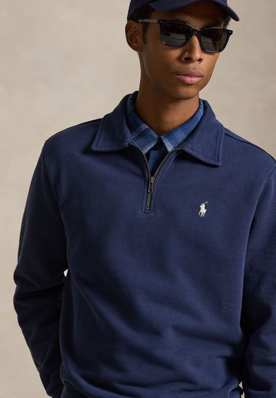 Mand iført mørkeblå kvartlyntrøje over blå ternet skjorte, mørke solbriller og navy baseballkasket, stående mod en neutral baggrund.