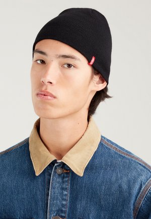 Black ribbed knit beanie with a red logo tag. Worn with a blue denim jacket featuring a contrasting tan corduroy collar and buttons.
