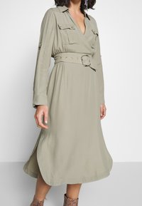 Khaki omslagklänning med långa ärmar, framfickor, en bälte med guldbeslag och en svepande A-linjeformad siluett. Slät tygtextur.