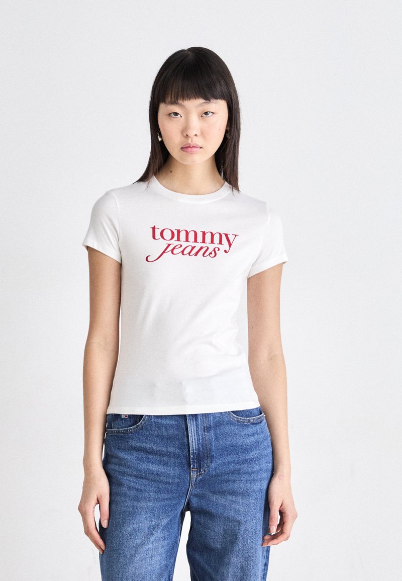 Balta medvilnės marškinėliai su raudonu „Tommy Jeans“ grafiniu spaudiniu priekyje. Prigludusi suknelė su trumpomis rankovėmis, derinama su mėlynomis džinsomis.