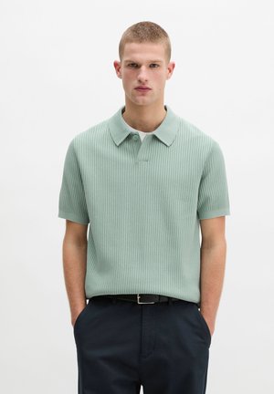 Mann trägt ein hellgrünes, geripptes Poloshirt mit kurzen Ärmeln, steckt es in schwarze Hosen mit schwarzem Gürtel und steht mit den Händen in den Taschen.